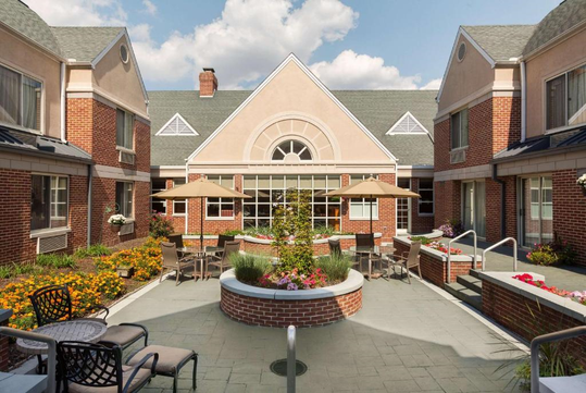 A courtyard with red brick buildings, tables with umbrellas, chairs, and flowerbeds under a partly cloudy sky.