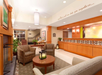 Hotel lobby with brown armchairs, a round table, and lantern centerpiece. Reception desk in the background, with artwork and plants decorating the area. Ceiling lights provide soft illumination.