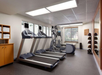 A small gym with treadmills, elliptical machine, exercise bike, towel rack, and medicine ball rack, located in a well-lit room with a window and wall mirror.