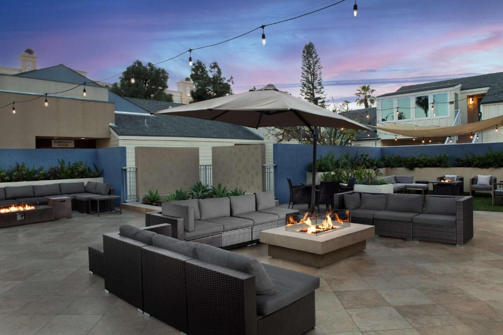 Outdoor patio area with gray wicker furniture around a square fire pit. String lights are overhead, and buildings are in the background against a sunset sky.
