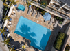 Aerial view of a rectangular swimming pool surrounded by lounge chairs, a small jacuzzi, an umbrella, and palm trees casting shadows on the poolside area.