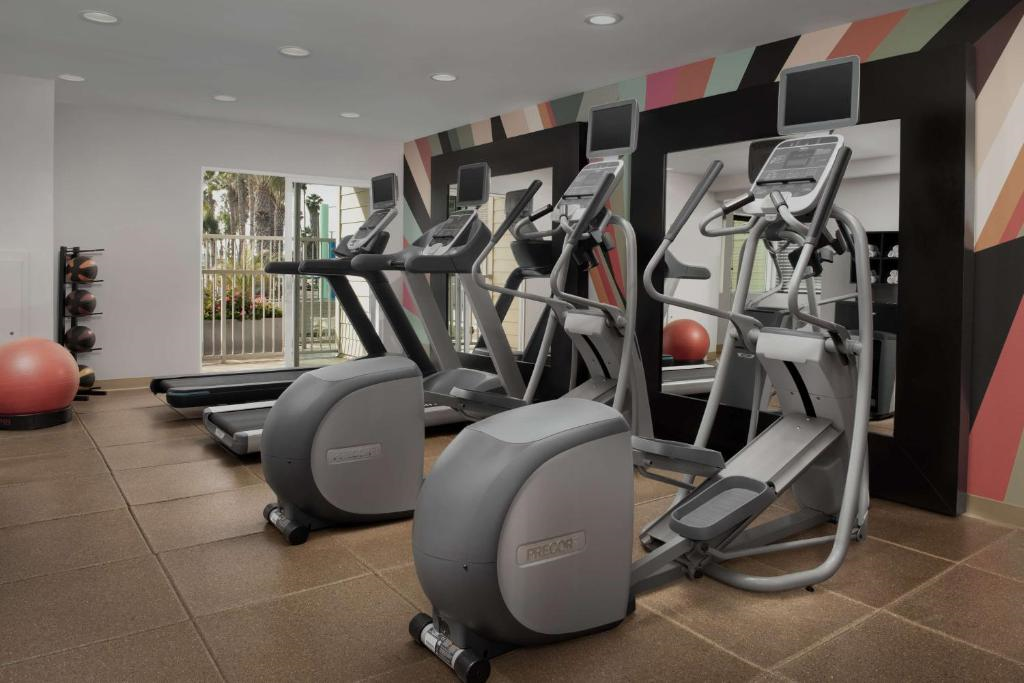 A modern gym with three elliptical machines, two treadmills, and a wall mirror. Exercise balls are visible in the corner. The wall is decorated with colorful geometric patterns.