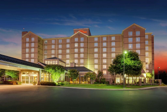 A large, multi-story hotel building is illuminated at dusk, with visible entrance canopy, landscaped greenery, and a clear, darkening sky in the background.