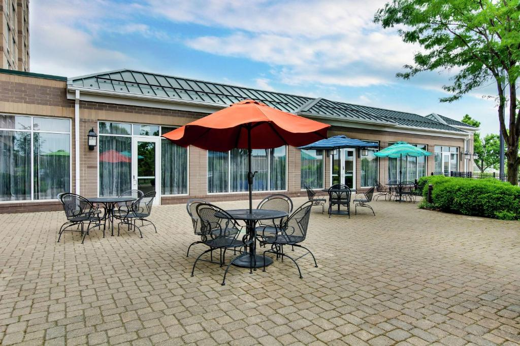 Outdoor patio with several metal tables and chairs, some shaded by large umbrellas, set on a paved surface next to a building with large windows.