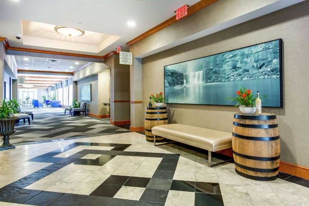 A hotel hallway with patterned tile floor, a beige bench, two barrel planters with flowers, framed waterfall art, and lounge seating visible in the distance.