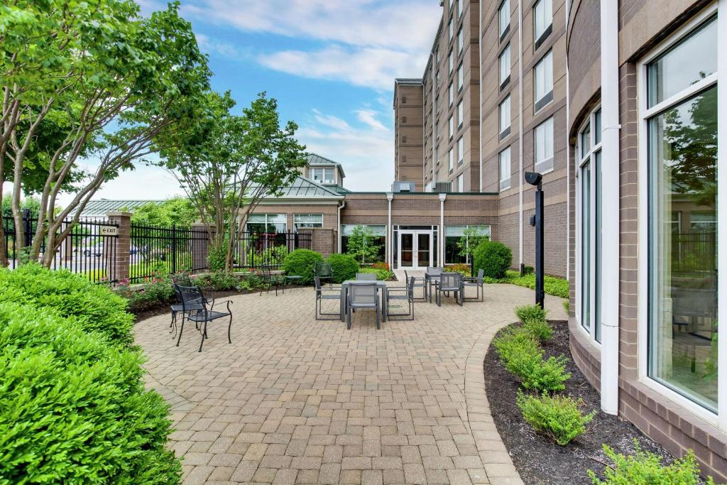 Brick patio with several tables and chairs, surrounded by green shrubs and trees, adjacent to a multi-story hotel building.