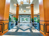 Modern hotel lobby with tall wooden columns, geometric tile flooring, hanging pendant lights, large windows, green plants, and seating areas near the entrance.