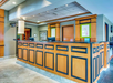 Hotel front desk area with wood paneling, two computer stations, overhead lighting, and framed artwork on the walls. Marble floor and neutral-toned decor are visible.