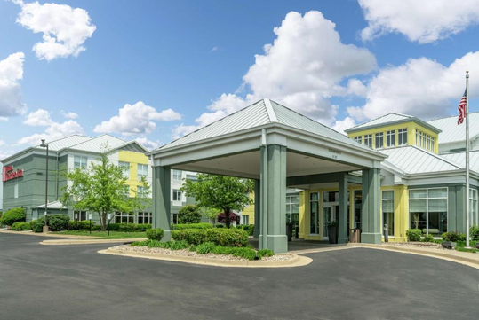A hotel building with a covered driveway entrance, surrounded by trees and landscaping under a partly cloudy sky.