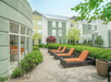 Outdoor patio area with wicker lounge chairs featuring orange cushions, surrounded by green bushes and trees, adjacent to a modern building with large windows.