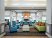 Modern hotel lobby with blue and green seating, a central ottoman, coffee tables, and a fireplace, surrounded by large windows with natural light.