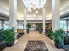 Spacious hotel lobby with tall white columns, modern hanging lights, large potted plants, tile flooring, and seating areas on both sides.