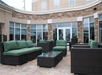 A stone terrace with several sets of green and gray patio furniture in front of a wall of windows at the Hilton Garden Inn New Braunfels.