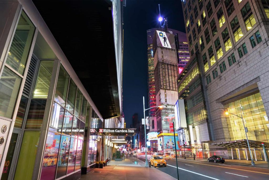 Night view of a city street with a Hilton Garden Inn sign, illuminated skyscrapers, billboards, and a yellow taxi on the street.