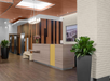 Modern hotel reception area with a wooden front desk, large plants, a cityscape photo on the wall, and pendant lighting.
