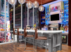 Modern bar with a sleek metal counter, brown chairs, and illuminated shelves stocked with bottles. Large windows offer a view of city buildings. Spherical pendant lights hang overhead.