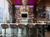 Modern bar interior with a sleek counter, six stools, and a variety of bottles on illuminated shelves. A flat-screen TV is centered above the bar.