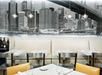 A modern restaurant interior with white tables, yellow and gray chairs, empty wine glasses, and a large black-and-white cityscape mural on the wall.
