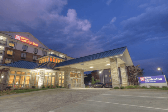 Exterior view of a Hilton Garden Inn hotel at dusk with a lit entrance and surrounding pathway.