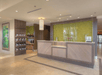 Hotel lobby with a wooden front desk, a green background, overhead lighting, and decorative shelves on the left. A painting is visible on the wall.