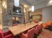 A meeting area with a wooden table, red chairs, and a lit stone fireplace. A TV is mounted above the fireplace. The room has modern lighting and wooden flooring.