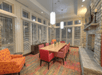 A dining room with a long wooden table, red and orange chairs, stone fireplace, and large windows with blinds. White pendant lights hang from the ceiling.