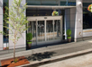 Street entrance with sliding glass doors, flanked by two potted plants and a small tree on the sidewalk. Hanging flower baskets are visible overhead.