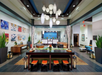 Bright, modern hotel lobby with a long table and orange stools, decorative plants, wall art, and hanging pendant lights. Reception desk in background.