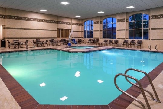 Indoor swimming pool with surrounding chairs and tables, metal handrails, large windows, and overhead lighting.