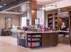 Hotel snack shop with shelves of drinks, snacks, and coffee machine. Wooden counters display assorted goods. Modern decor with seating area in the background.