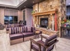 Modern lounge area with stone fireplace, purple cushioned seating, armchair, and side table. Large windows and a vase with flowers on a console table add touches of decor.