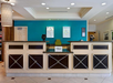 Hotel reception area with a front desk, turquoise accent wall, framed artworks, light fixtures, and digital check-in screens. A Hilton sign is on the counter.