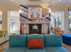 Modern living room with a teal sofa, orange cushion, and a geometric-patterned fireplace. Above the fireplace is a colorful abstract painting. Neutral walls and large windows frame the space.