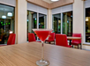 A red cocktail in a martini glass sits on a wooden table in a restaurant with red chairs and large windows in the background.