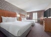A hotel room with two double beds, a desk with a chair, a flat-screen TV, and a window with curtains. Walls have patterned wallpaper and carpeted floor.