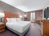 Hotel room with a king-sized bed, two bedside tables with lamps, an armchair, a work desk and chair, a flat-screen TV on a dresser, and patterned curtains.