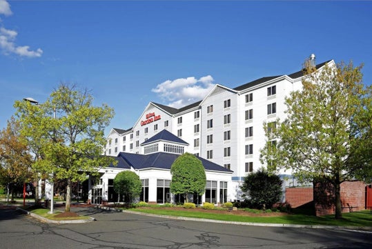 Exterior view of a Hilton Garden Inn with a blue roof, surrounded by trees and greenery under a clear sky.