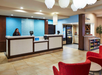 Hotel reception area with a front desk, a staff member, seating on the right, a snack vending area, and hanging lights above.