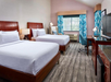 Hotel room with two double beds, wooden furniture, a desk, chair, TV, and window with blue patterned curtains. Neutral-toned walls and carpeted floor.