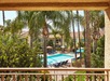 View from a balcony overlooking a palm tree-lined pool area with lounge chairs and umbrellas, set against a backdrop of a stucco building with a red-tiled roof.