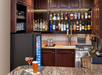 A bar with a granite counter, featuring a variety of liquor bottles on shelves, a draft beer tap, a small fridge, and a cocktail and glass of beer on the counter.
