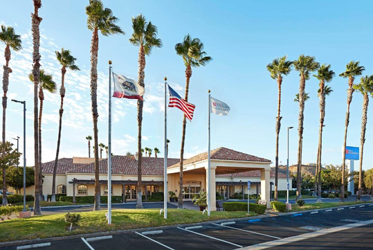 A building with a tiled roof is surrounded by palm trees. Three flags are flying in front: California, United States, and a corporate flag. An adjacent blue sign is visible.