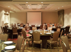 A banquet room set up with round tables and chairs, ready for an event. Plates, glasses, and napkins are arranged on the tables. A projection screen is visible in the background.
