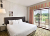 Hotel room with a large bed, bedside tables, and a sliding glass door leading to a balcony with chairs, overlooking greenery.
