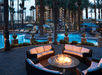 A patio with a fire pit with couches around it and a luxurious tropical swimming pool with bright blue water and palm trees in the background.