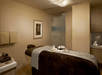 A spa room with a massage table with towels and stones layed out on it, a shower on the back wall, and a cart of towels on the left.