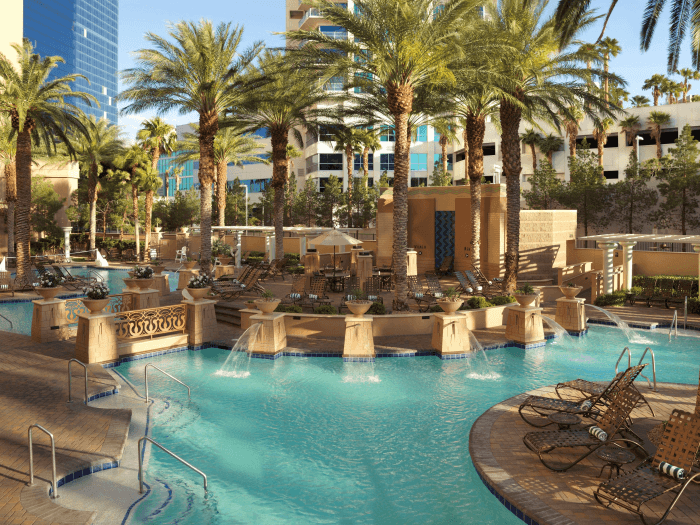 A luxurious tropical outdoor swimming pool with lounge chairs and palm trees around it and hotels in the background.