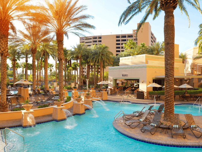 A luxurious tropical outdoor swimming pool with islands in the middle with palm trees and lounge chairs with a restaurant and hotel in the background.