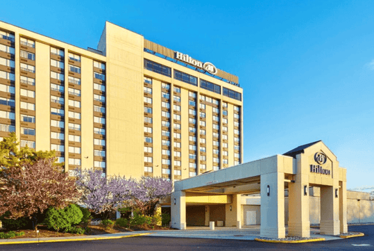 A tall, multi-story Hilton hotel building with a prominent logo, surrounded by trees and a clear sky. The entrance features a covered driveway.