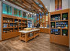 A modern convenience store interior with wooden shelves stocked with snacks, drinks, and various items. A central display table holds more products. Bright lighting and colorful ceiling panels.